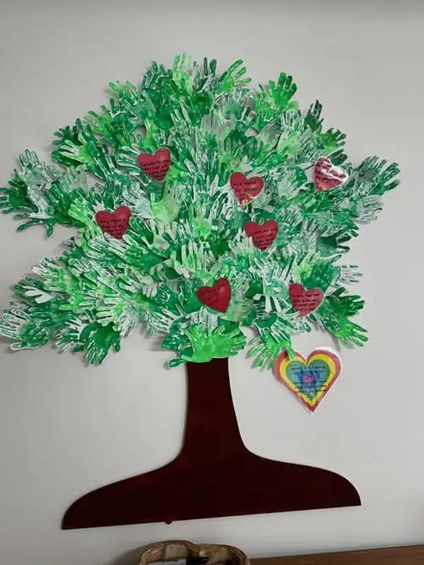 Kindness Tree: a wooden tree on the wall with green handprints and red paper hearts noting acts of kindness.