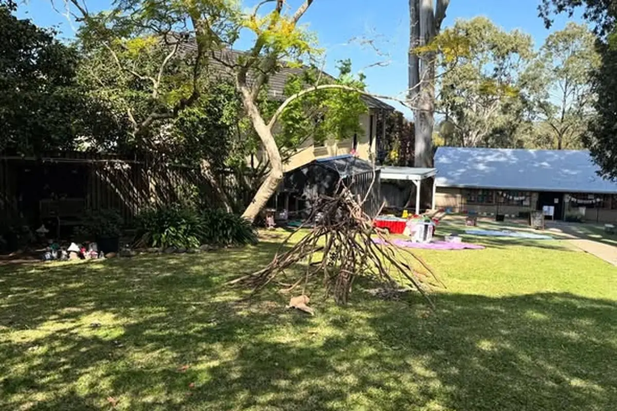 Open playground with a teepee-shaped branch structure under gum trees, with children's learning materials set out on rugs.